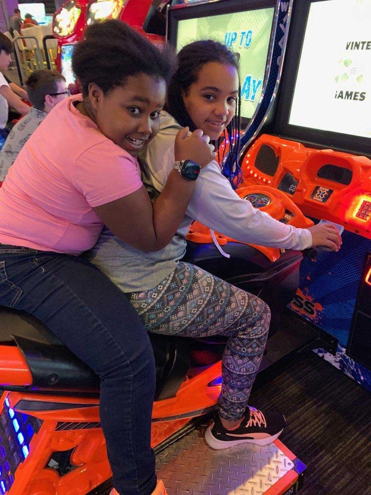 Two girls sharing the seat of a motorcycle arcade game, turning to smile at the camera.