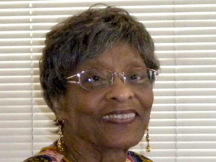 Portrait of a smiling older woman wearing glasses and drop earrings, photographed indoors in front of window blinds.