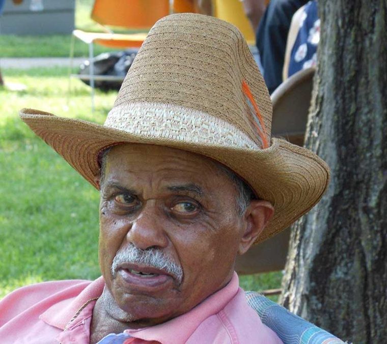 An older man in a straw hat and pink polo shirt seated under a tree at an outdoor family gathering.