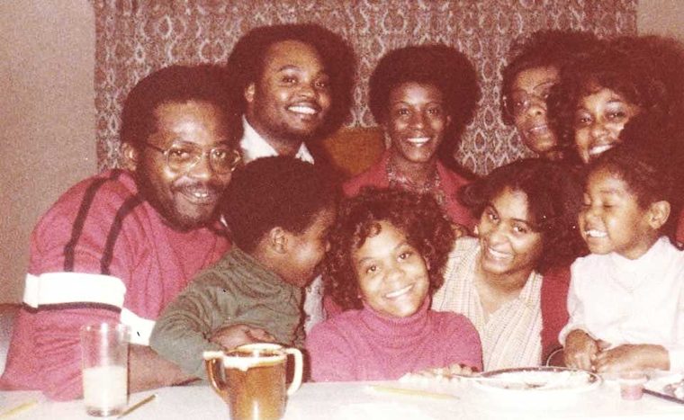 A vintage photograph of nine family members of several generations crowded together and smiling around a table set with cups and plates.