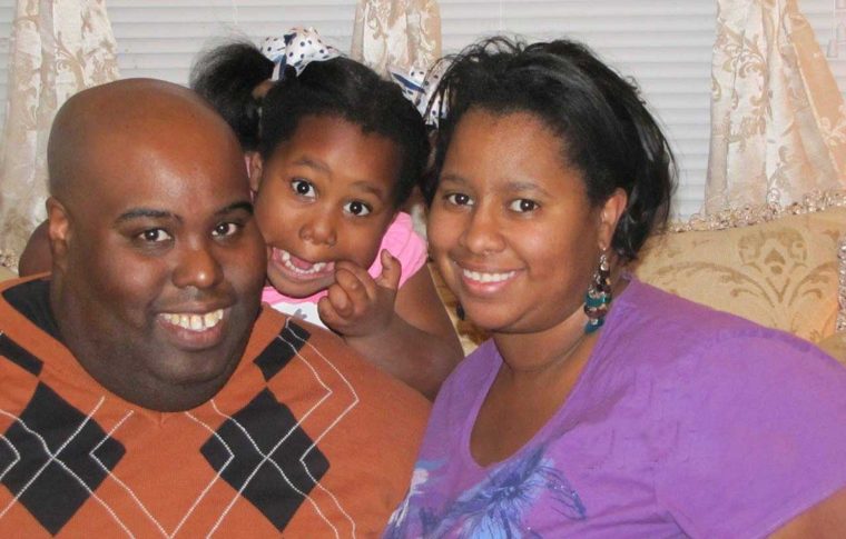 Two smiling adults on a sofa with a young child in a hair bow grinning between them.